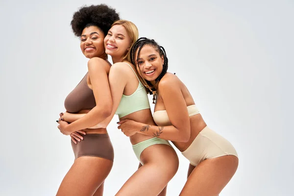 Jovem mulher americana africana alegre em lingerie abraçando amigos e olhando para a câmera enquanto posando isolado em cinza, diferentes tipos de corpo e conceito de auto-aceitação, modelos multiculturais — Fotografia de Stock