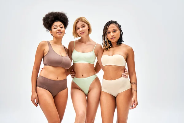 Young and blonde woman in lingerie hugging african american friends and looking at camera while standing isolated on grey, different body types and self-acceptance concept, multicultural models — Stock Photo