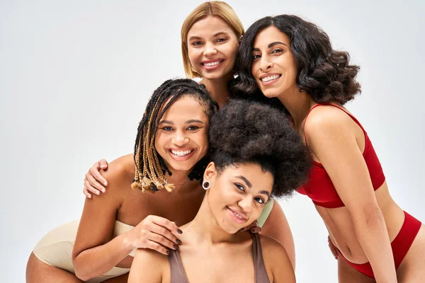 Femmes multiethniques positives en lingerie colorée et moderne regardant la caméra tout en posant ensemble isolé sur gris, différents types de corps et concept d'acceptation de soi, modèles multiculturels — Photo de stock