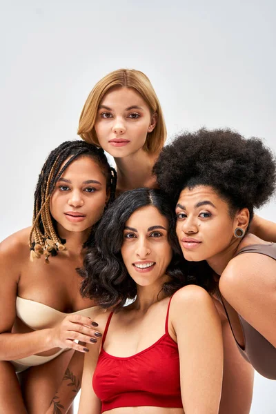 Mulheres multiétnicas positivas em sutiãs modernos e coloridos olhando para a câmera juntos enquanto posando isolado em cinza, diferentes tipos de corpo e conceito de auto-aceitação, modelos multiculturais — Fotografia de Stock