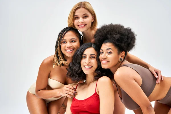 Портрет группы улыбающихся мультикультурных женщин в красочных бюстгальтерах, смотрящих в камеру и обнимающих изолированных на сером, разных типах тела и концепции самовосприятия, мультикультурных моделях — стоковое фото