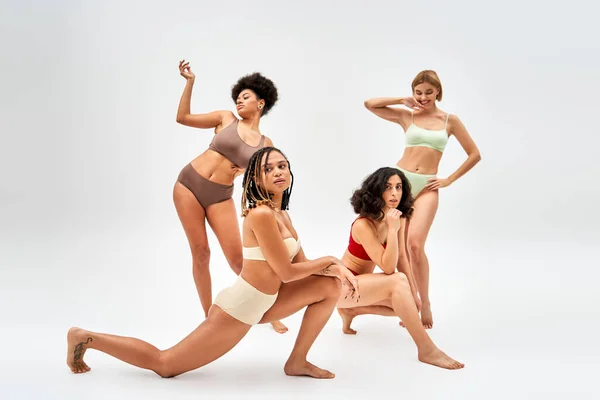 Pleine longueur de femme afro-américaine tatouée en lingerie moderne posant à côté de copines multiethniques debout sur fond gris, différents types de corps et concept d'acceptation de soi — Photo de stock