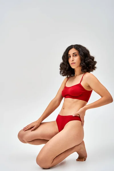 Full length of sexy middle eastern brunette woman in red lingerie holding hand on hip and looking at camera while posing on grey background, self-acceptance and body positive concept — Stock Photo
