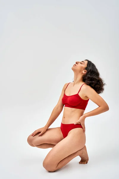 Side view of middle eastern brunette woman in modern red lingerie holding hand on hip and posing with closed eye on grey background, self-acceptance and body positive concept — Stock Photo