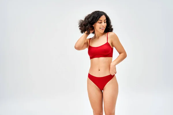 Smiling and brunette middle eastern model in red bra and panties touching hair and looking away while standing and posing isolated on grey, body acceptance concept — Stock Photo