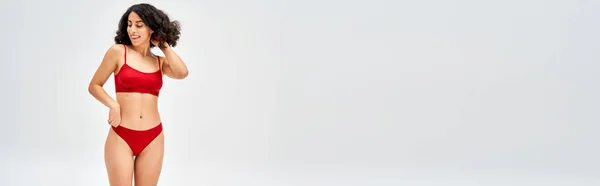 Femme positive du Moyen-Orient en soutien-gorge rouge et culotte touchant les cheveux et regardant loin tout en se tenant debout et posant isolé sur gris, concept d'acceptation du corps avec espace de copie, bannière — Photo de stock
