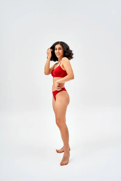 Full length of barefoot and middle eastern woman in modern red lingerie touching hair and looking at camera while standing on grey background, self-acceptance and body positive concept — Stock Photo