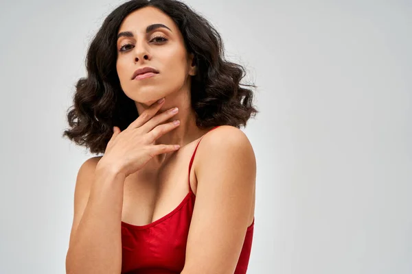 Portrait of brunette middle eastern woman in trendy red bra touching neck and looking at camera while standing isolated on grey, self-acceptance and body positive concept — Stock Photo