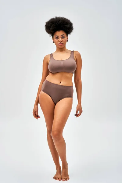 Comprimento total de mulher americana africana confiante e descalça em lingerie marrom olhando para a câmera enquanto em pé e posando em fundo cinza, auto-aceitação e conceito positivo para o corpo — Fotografia de Stock