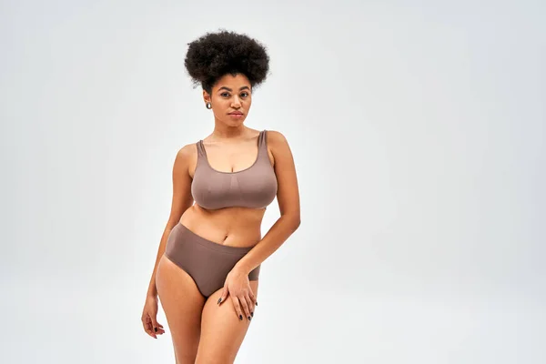 Confident african american woman in modern brown bra and panties looking at camera while touching hip and posing isolated on grey with copy space, self-acceptance and body positive concept — Stock Photo