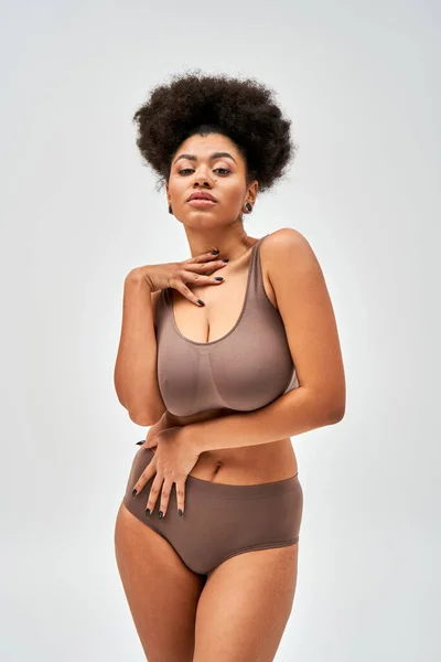 Portrait de séduisante femme afro-américaine en lingerie marron moderne touchant le cou et regardant la caméra tout en restant isolée sur le concept gris, auto-acceptation et corps positif — Photo de stock