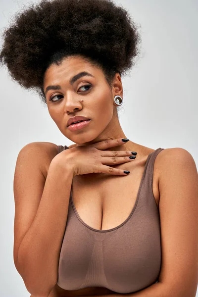 Retrato de mujer afroamericana segura y sexy en sujetador marrón tocando el cuello y mirando hacia otro lado mientras está de pie y posando aislado en gris, auto-aceptación y el concepto de cuerpo positivo - foto de stock