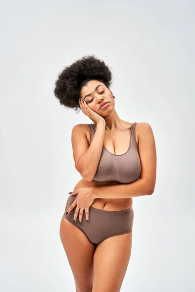 Mulher americana africana sedutora em lingerie marrom moderno tocando bochecha enquanto posando com os olhos fechados e de pé isolado em cinza, auto-aceitação e corpo conceito positivo — Fotografia de Stock
