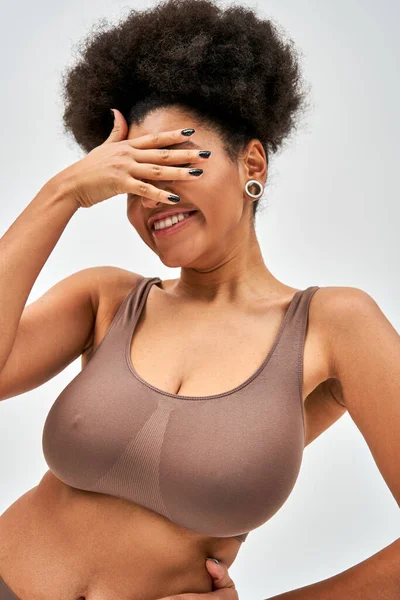 Mulher afro-americana alegre em sutiã marrom moderno cobrindo rosto com a mão enquanto em pé e posando isolado em cinza, auto-aceitação e corpo conceito positivo — Fotografia de Stock