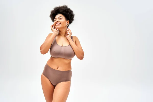 Femme afro-américaine positive et sexy en lingerie brune moderne touchant la joue et détournant les yeux tout en se tenant debout et posant isolé sur le gris, l'acceptation de soi et le corps concept positif — Photo de stock