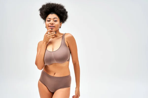 Positive and seductive african american woman in brown panties and bra touching chin and looking at camera while standing  isolated on grey, self-acceptance and body positive concept — Stock Photo