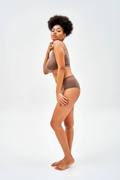 Full length of barefoot and sexy african american model in modern brown lingerie touching shoulder and looking at camera on grey background, self-acceptance and body positive concept — Stock Photo
