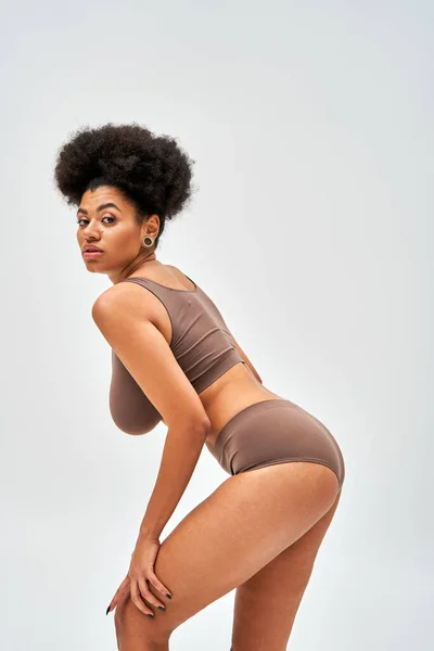 Femme afro-américaine confiante et moderne en lingerie marron regardant la caméra tout en touchant la jambe et posant isolée sur le concept gris, l'acceptation de soi et le corps positif — Photo de stock