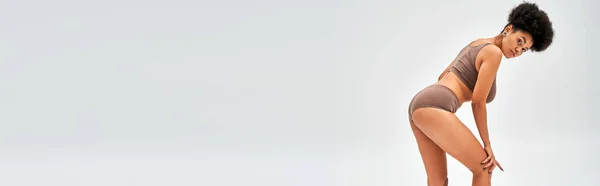 Confiante mulher afro-americana em calcinha marrom e sutiã tocando joelho e olhando para a câmera enquanto posando isolado em cinza, auto-aceitação e corpo conceito positivo, banner com espaço de cópia — Fotografia de Stock