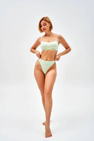 Full length of barefoot and blonde young woman in light green lingerie touching panties while standing and posing on grey background, self-acceptance and body positive concept — Stock Photo