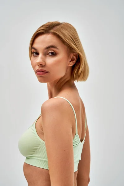 Porträt einer selbstbewussten jungen blonden Frau mit natürlichem Make-up, die einen hellgrünen BH trägt und in die Kamera blickt, während sie isoliert auf Grau, Selbstakzeptanz und einem körperbetonten Konzept steht — Stockfoto