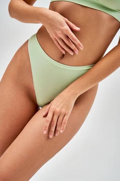 Cropped view of seductive young woman in light green panties and bra touching belly and leg while standing isolated on grey, self-acceptance and body positive concept, sensuality, femininity — Stock Photo