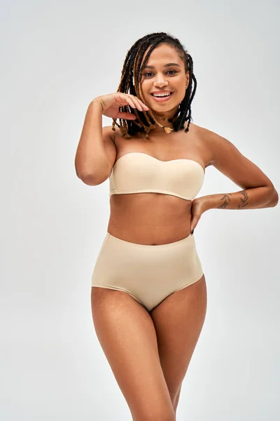 Femme afro-américaine joyeuse et tatouée en lingerie beige tenant la main sur la hanche et regardant la caméra tout en restant isolée sur le concept gris, auto-acceptation et corps positif — Photo de stock