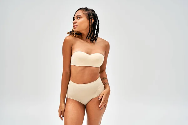 Modern young african american woman with natural makeup wearing beige bra and panties while touching hip isolated on grey, self-acceptance and body positive concept, femininity — Stock Photo