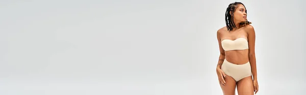 Moderno jovem modelo afro-americano com maquiagem natural vestindo lingerie bege sexy e tocando quadril isolado em cinza, auto-aceitação e conceito de corpo positivo, banner com espaço de cópia — Fotografia de Stock