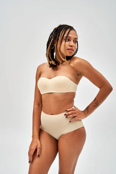 Femme afro-américaine confiante tatouée et jeune avec un maquillage naturel portant une lingerie beige et posant tout en étant isolée sur un concept gris, d'acceptation de soi et positif pour le corps — Photo de stock