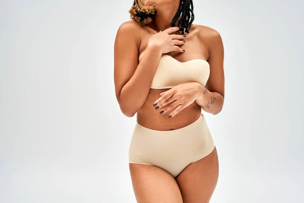 Vue recadrée de jeune femme afro-américaine tatouée en soutien-gorge beige et culotte touchant la poitrine tout en se tenant isolé sur gris, acceptation de soi et concept positif du corps — Photo de stock