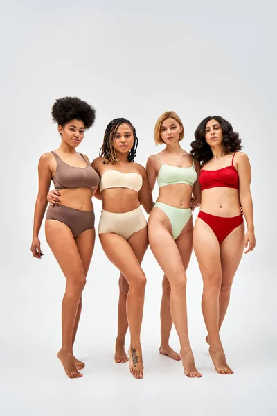 Comprimento total de mulheres multiétnicas sexy e descalça em lingerie colorida abraçando e olhando para a câmera em fundo cinza, modelos multiculturais e conceito de movimento de positividade corporal — Fotografia de Stock