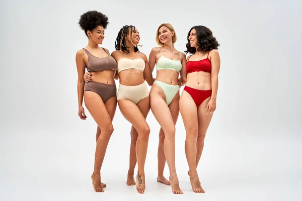 Barefoot and multiethnic girlfriends in colorful modern lingerie hugging and smiling at each other on grey background, multicultural models and body positivity movement concept — Stock Photo