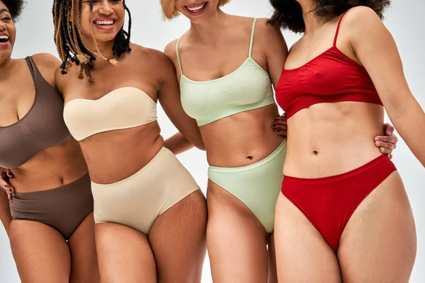 Cropped view of cheerful multiethnic women in colorful and modern lingerie standing and hugging each other isolated on grey, multicultural models and body positivity movement concept — Stock Photo