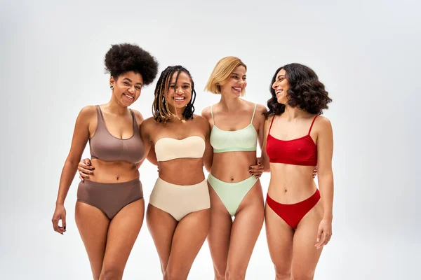 Cheerful multiethnic girlfriends with natural makeup hugging while posing in colorful lingerie and standing isolated on grey, multicultural models and body positivity movement concept — Stock Photo