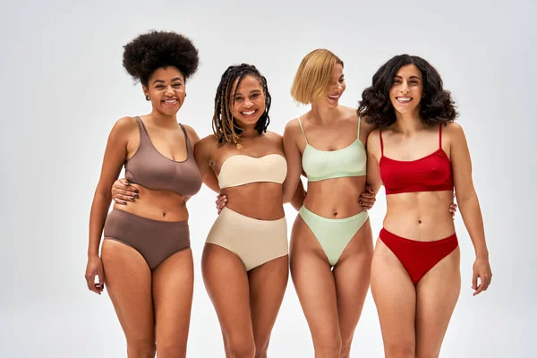 Cheerful and sexy multiethnic women in colorful lingerie hugging and looking at camera together isolated on grey, different body types and self-acceptance, multicultural representation — Stock Photo