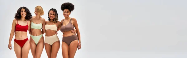 Laughing and confident multiethnic women in colorful lingerie hugging and posing together isolated on grey, different body types and self-acceptance, multicultural representation, banner — Stock Photo