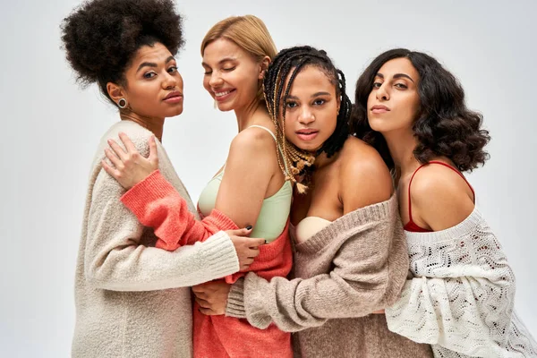 Portrait of group of multiethnic girlfriends in warm knitted jumpers hugging and standing together isolated on grey, different body types and self-acceptance, multicultural representation — Stock Photo