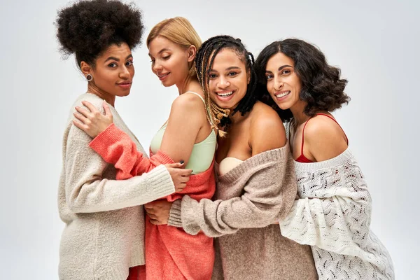 Весёлые многоэтнические женщины, обнимающие своих подружек в шерстяных вязаных свитерах и стоящие изолированные на сером, разных типах тела и принятии себя, мультикультурном представлении — стоковое фото