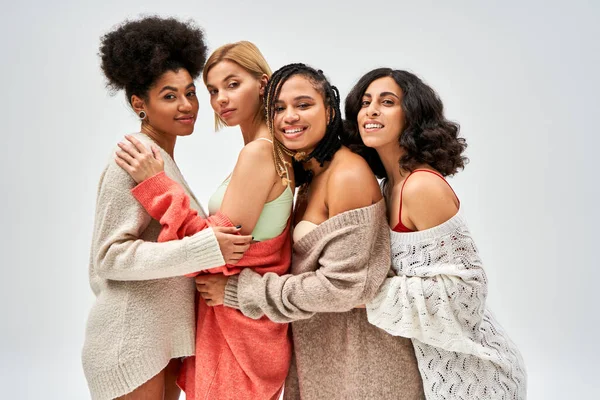 Retrato de mujeres multiétnicas con estilo y confianza en suéteres de punto sonriendo y abrazándose aislados en gris, diferentes tipos de cuerpo y auto-aceptación, representación multicultural - foto de stock