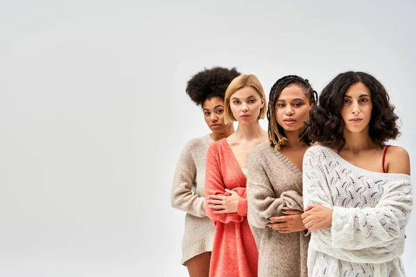 Grupo multiétnico de mujeres en suéteres de punto elegantes cruzando los brazos y mirando a la cámara aislada en gris, diferentes tipos de cuerpo y auto-aceptación, representación multicultural, espacio de copia - foto de stock
