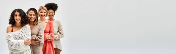 Positive und multiethnische Freundinnen in stylischen warmen Pullovern, die die Arme verschränken, während sie isoliert auf grauen, unterschiedlichen Körpertypen und Selbstakzeptanz posieren und stehen, multikulturelle Repräsentation, Banner — Stockfoto