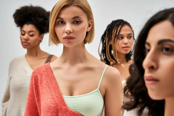 Portrait of young blonde woman in bra and wool jumper looking at camera while standing near blurred friends isolated on grey, different body types and self-acceptance, multicultural representation — Stock Photo