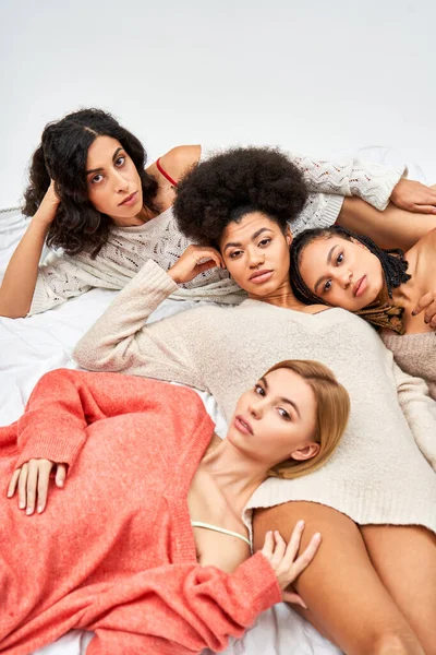High angle view of multiethnic women in cozy knitted jumpers looking at camera while lying on bed isolated on grey, different body types and self-acceptance, multicultural representation — Stock Photo
