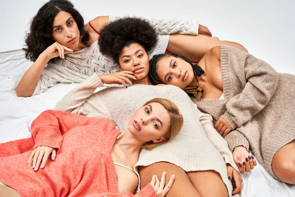Високий кут зору на багатоетнічну групу жінок у стильних теплих стрибках, які дивляться на камеру, лежачи на ліжку ізольовано на сірому, різних типах тіла та самоприйнятті, мультикультурне представлення — стокове фото