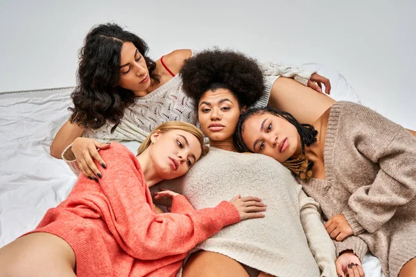 Vista de ángulo alto de las mujeres multiétnicas en elegantes jerséis cálidos mirando a la cámara mientras yacen en la cama aislados en gris, diferentes tipos de cuerpo y auto-aceptación, representación multicultural - foto de stock