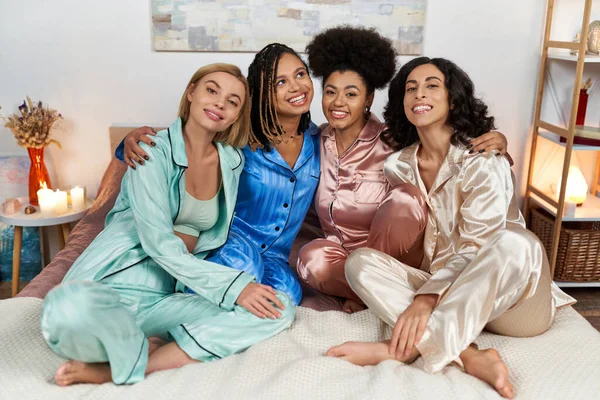 Веселые и многонациональные девушки в красочных пижамах, обнимающие и смотрящие в камеру, сидя на кровати во время пижамной вечеринки дома, проводя время в удобной сонной одежде, пижамной вечеринке — стоковое фото