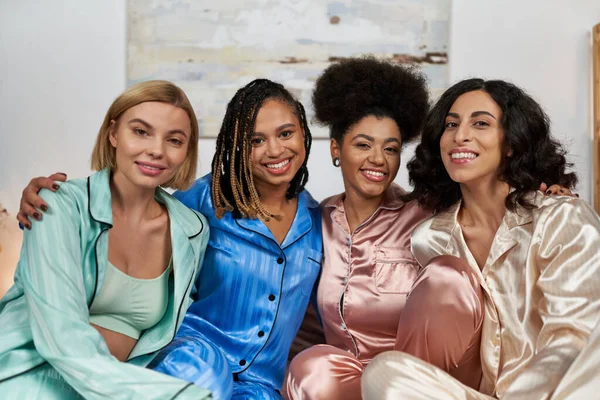 Portrait de femmes afro-américaines positives embrassant des copines multiethniques en pyjama coloré et regardant la caméra pendant la soirée pyjama, collant le temps dans des vêtements de nuit confortables, soirée pyjama — Photo de stock