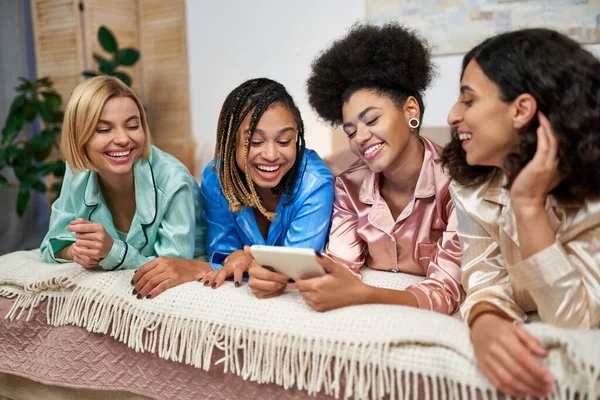 Smiling african american woman in modern pajama holding smartphone near multiethnic girlfriends lying and relaxing on bed during pajama party at home, bonding time in comfortable sleepwear — Stock Photo