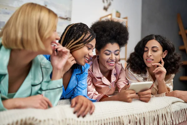Positive afrikanisch-amerikanische Frau hält Smartphone in der Nähe verschwommener multiethnischer Freundinnen im bunten Pyjama auf dem Bett liegend während Pyjama-Party im Schlafzimmer zu Hause, Bindungszeit in bequemer Nachtwäsche — Stockfoto